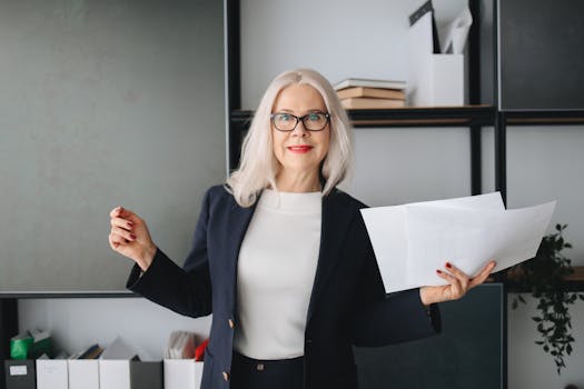 Elderly woman in a black blazer, holding papers while smiling indoors, epitomizing confidence and professionalism.